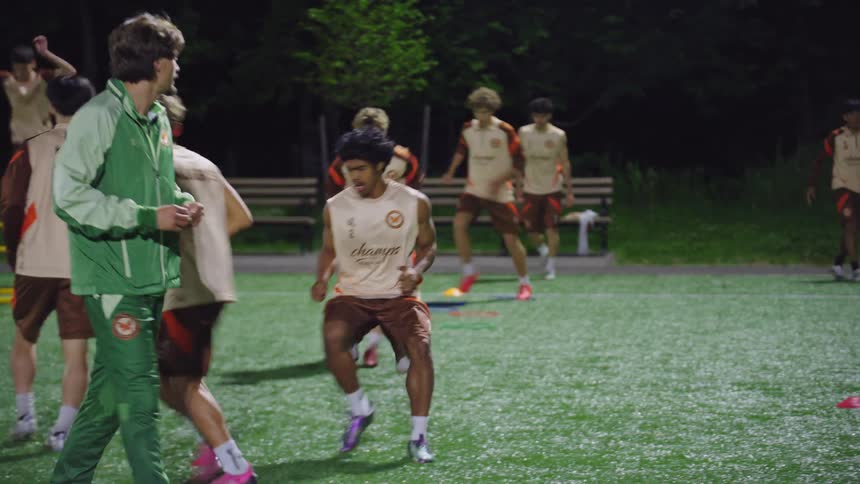 A soccer team in matching uniforms performs agility drills on a turf field at night while their coach watches in slow motion