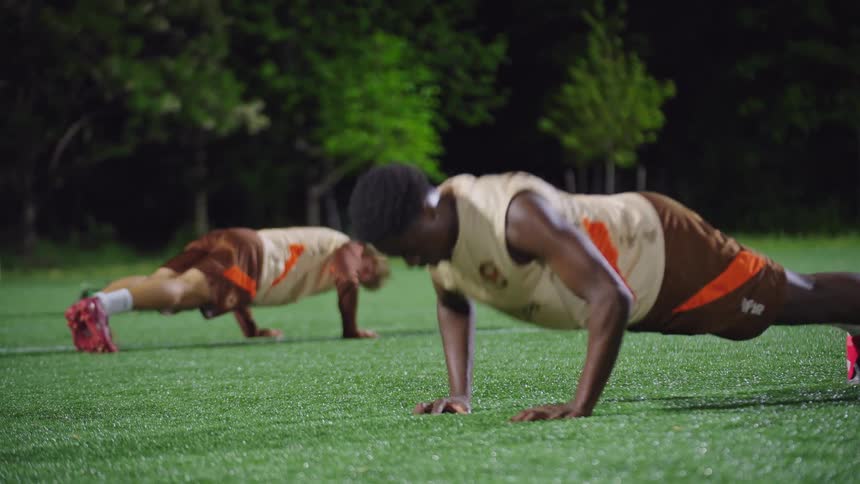Two diverse male athletes do push-ups together on an artificial turf field during a nighttime workout session