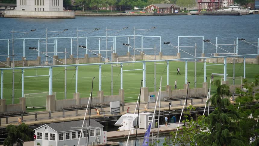 An elevated, long shot of a waterside soccer field where a few people are playing next to a marina