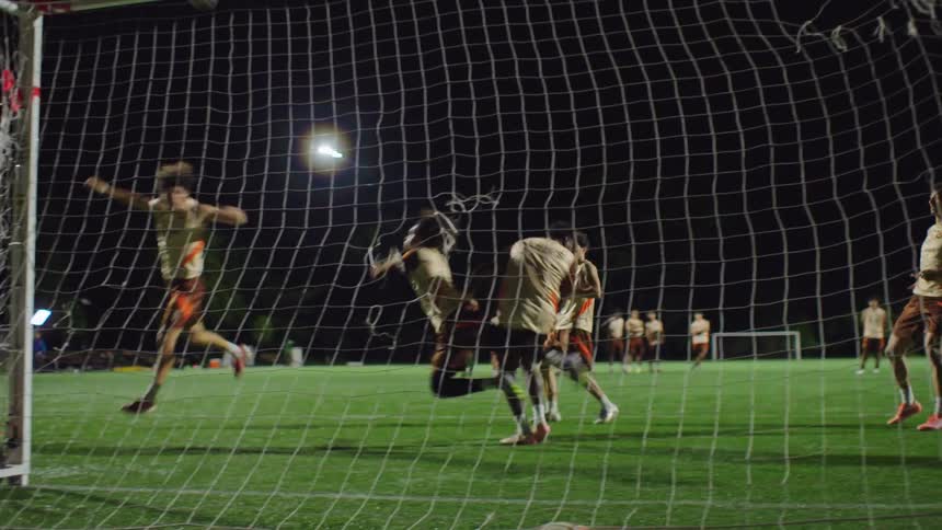 Slow motion shot from behind the goal as a soccer player scores a penalty kick past the diving goalie during a nighttime practice session