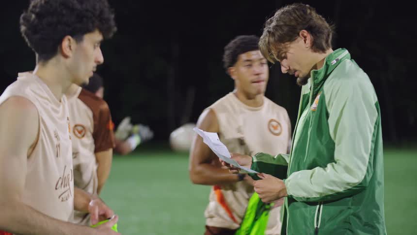 A dedicated soccer coach instructs his multicultural team on game strategy during a night training session on the field