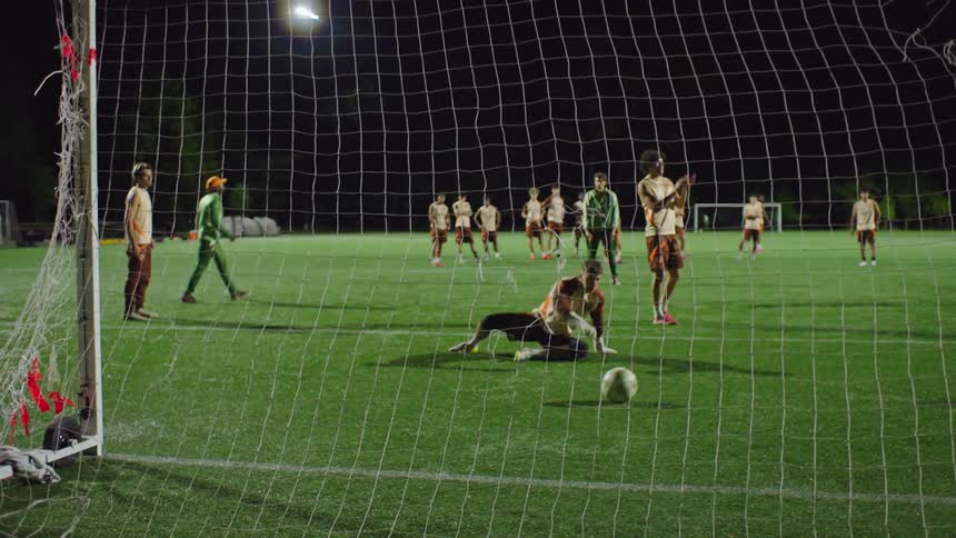 Slow motion view from behind the goal of a soccer team practicing penalty kicks at night, where the goalie makes a great save and then gets scored on