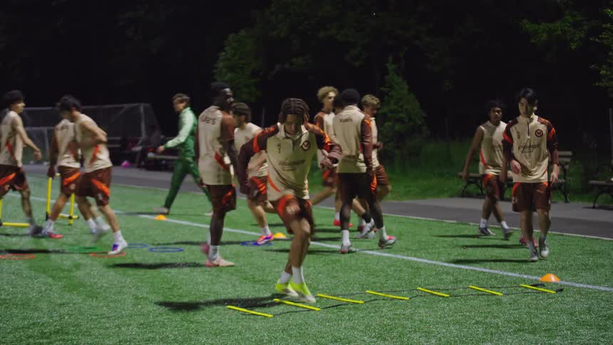 In slow motion, a team of diverse soccer players diligently practices footwork and agility drills on a green turf field at night