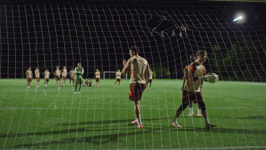 From behind the goal net, a soccer player scores with a header in slow motion during a night match as the team celebrates