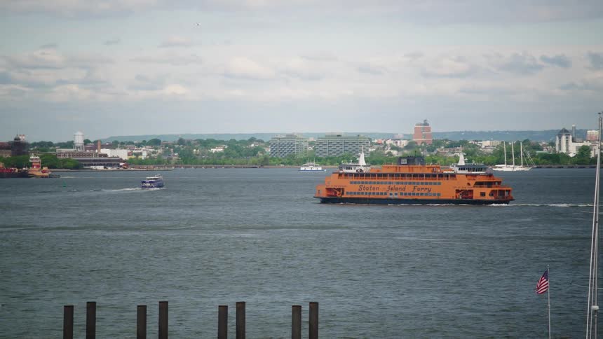 The iconic orange Staten Island Ferry sails across the river with the distant cityscape in the background on a cloudy day