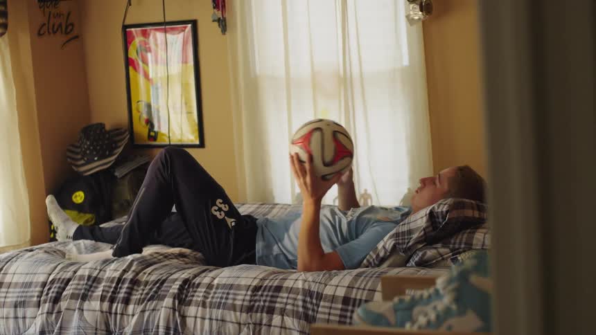 A relaxed teenage athlete lies on their bed in a sunlit room, casually playing with a soccer ball