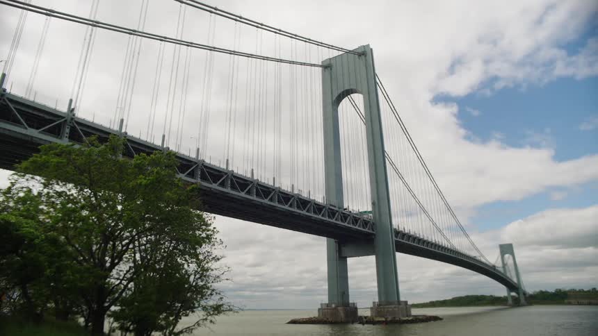 A static, low-angle shot of the Verrazzano-Narrows Bridge spanning the water on a partly cloudy day in New York City