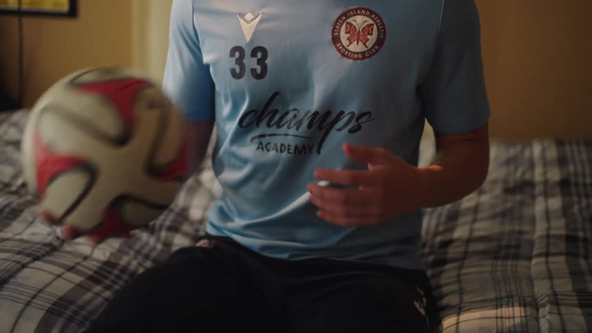 A contemplative teenage soccer player in his team jersey sits on his bed and spins a ball in his hands