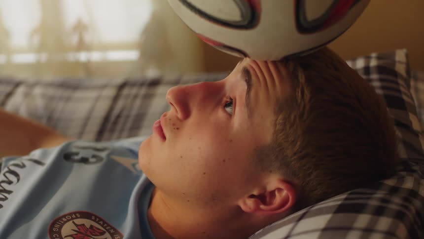 A focused young soccer player in a jersey concentrates on balancing a soccer ball on his forehead while lying in his bed