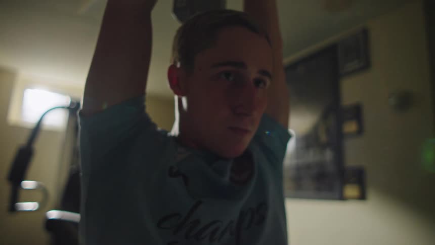 A focused teenage boy lifts a dumbbell over his head while working out in his home gym