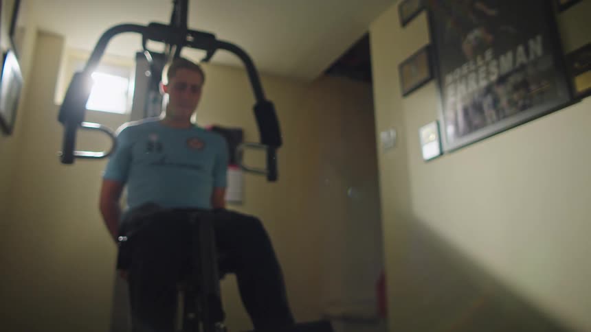 A low-angle slow-motion shot shows a male athlete in his cleats working out his legs on a home gym machine