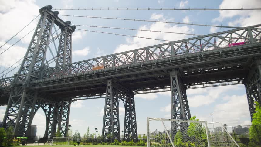 A low-angle shot tilts down from the steel trusses of the Williamsburg Bridge to a bright green soccer field in a park below