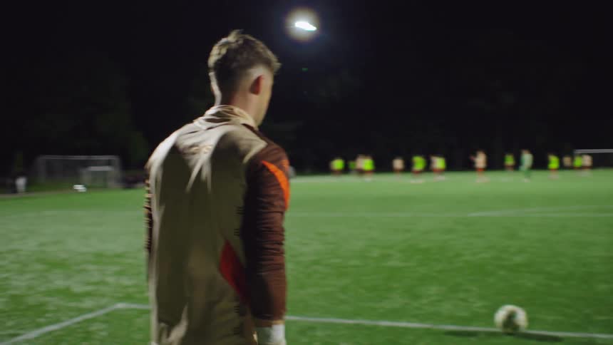 A soccer goalkeeper walks to the line, places the ball, and kicks it downfield during a night practice session