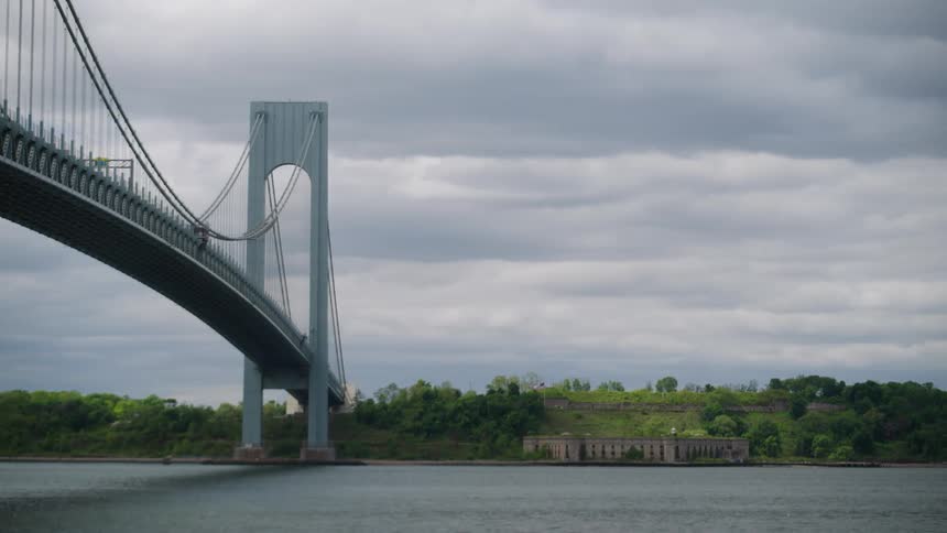 A wide, static shot shows the iconic Verrazzano-Narrows Bridge crossing over the bay towards Fort Wadsworth on a cloudy day