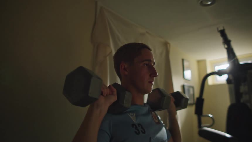 A determined young man performs dumbbell shoulder presses in a dimly lit home gym in slow motion