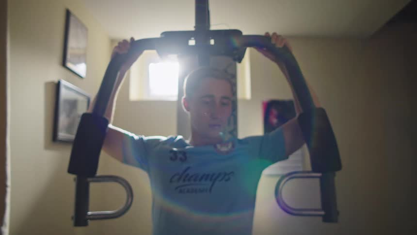Slow motion dolly-out shot reveals a young man intensely working out on a home gym machine with dramatic backlighting