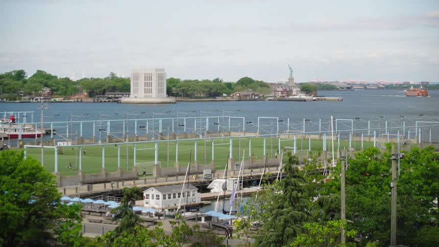 A scenic view of New York Harbor showing a waterfront athletic field, with Governors Island and the Statue of Liberty in the distance