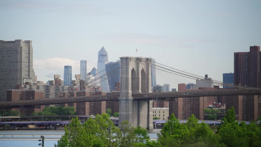 A static, wide shot shows the iconic Brooklyn Bridge with the New York City skyline in the background on a hazy day