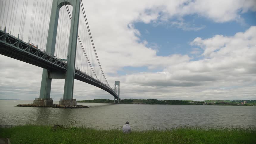 A lone person sits on the grassy riverbank contemplating the view of the massive Verrazzano Bridge in New York City