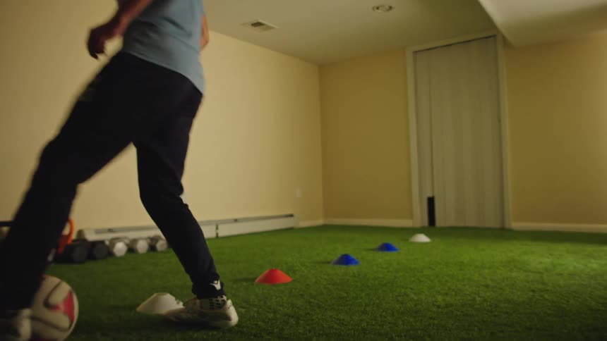 A young male athlete practices soccer dribbling drills in slow motion around cones on an indoor turf field