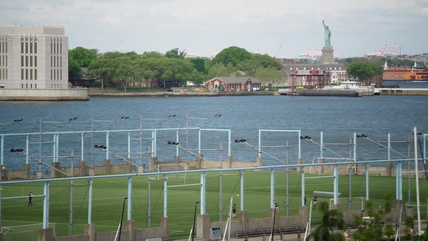 A pan right shot across the East River in New York City reveals the Statue of Liberty and a passing Staten Island Ferry behind a waterside soccer field