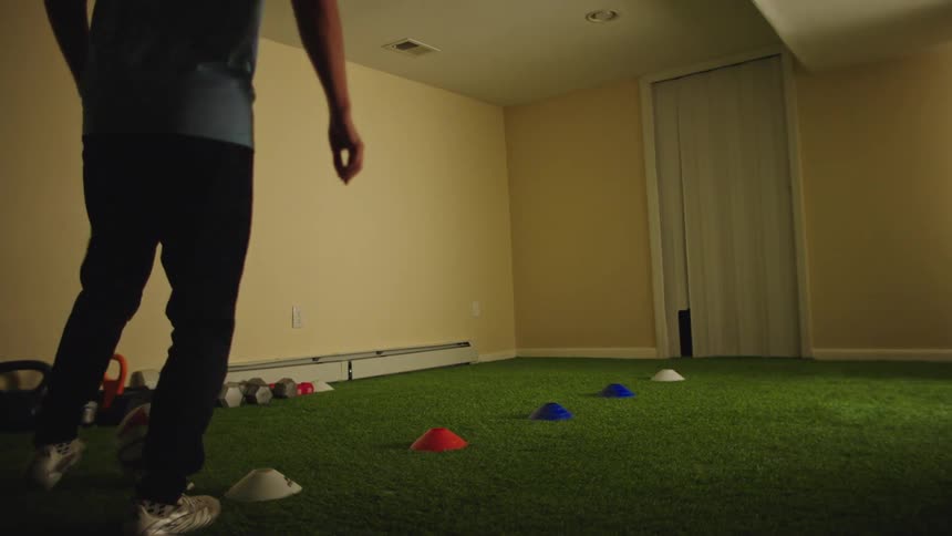 A young man practices his soccer skills, dribbling a ball around cones on an indoor turf field in his home