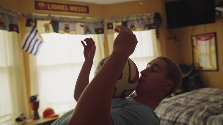A talented young soccer player practices juggling and balancing a ball in slow motion inside his bedroom