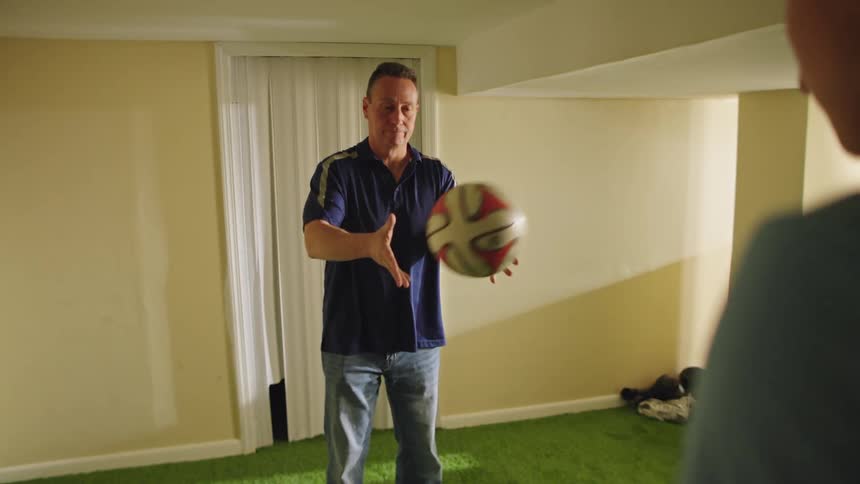 A father and son toss a soccer ball back and forth in slow motion while standing on a green turf floor in their basement