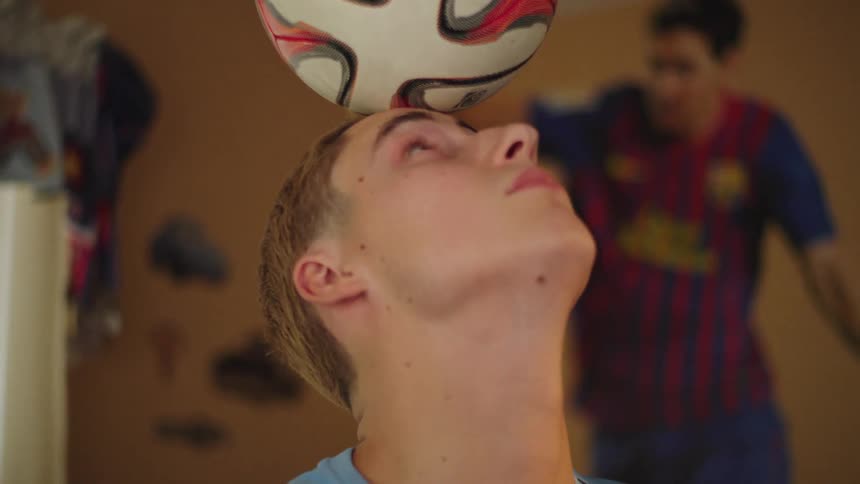 A young soccer player in his bedroom practices his skills by balancing a soccer ball on his forehead in slow motion