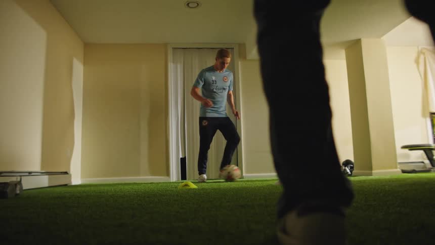 Two male soccer players pass a ball back and forth while practicing footwork drills in slow motion on an indoor turf field