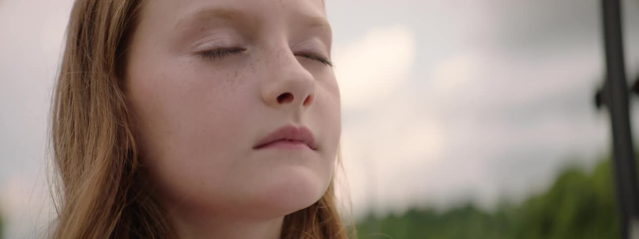 A thoughtful young girl with red hair and freckles closes her eyes in quiet contemplation outdoors