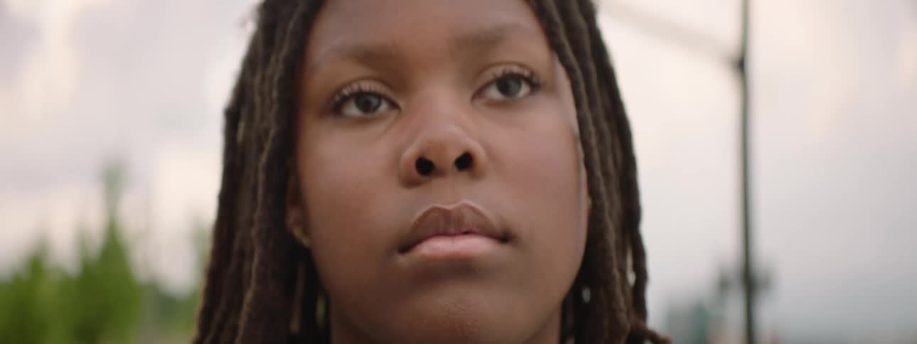 A slow rack focus reveals a close-up portrait of a serious young person with dreadlocks looking thoughtfully into the distance