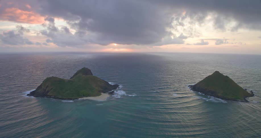 A serene wide shot of two tropical islands in the vast ocean as the sun rises on the horizon under a cloudy sky