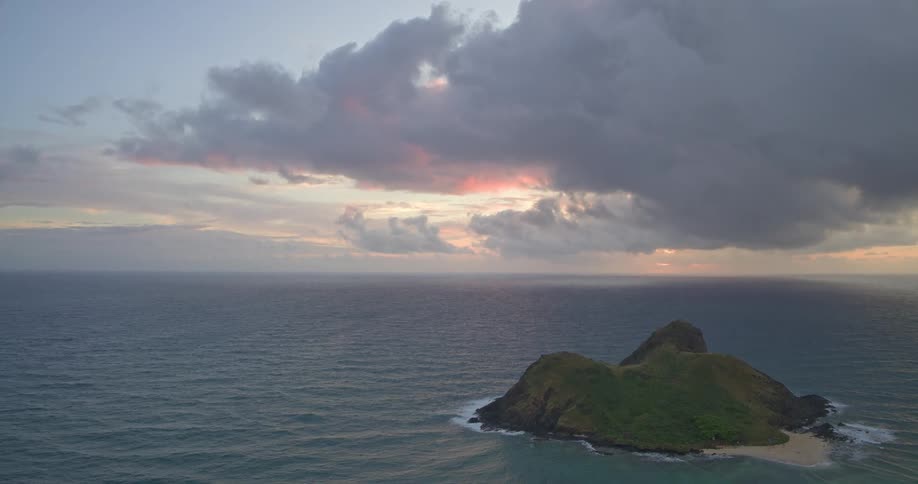 A wide, high-angle view captures a secluded tropical island in the ocean under a dramatic cloudy sky at sunset