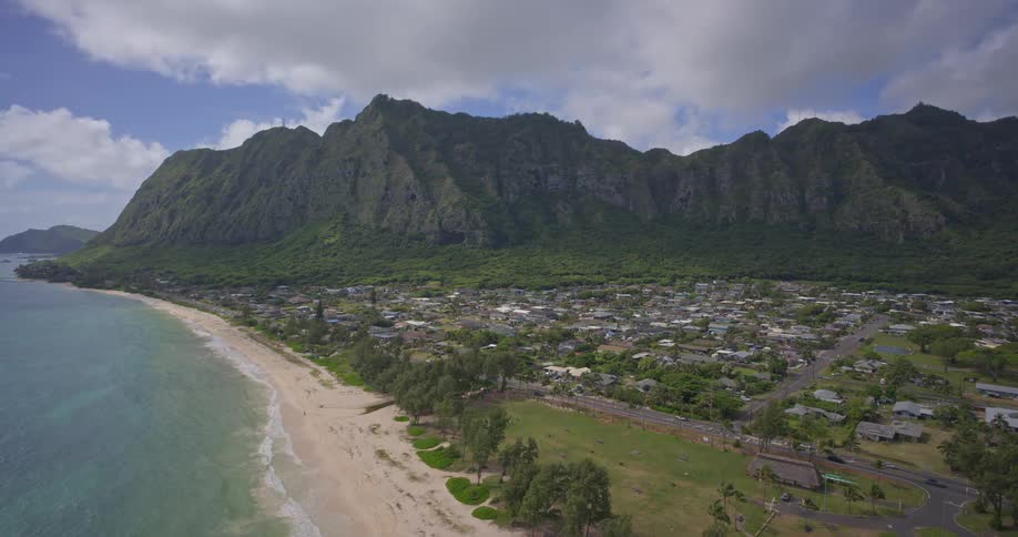 A wide aerial shot captures the stunning Hawaiian coastline, where a small town is nestled between a sandy beach and dramatic, lush green mountains