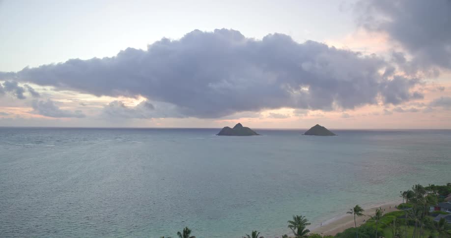 An aerial drone shot flies over a tropical coastline with palm trees and out to the ocean towards two small islands during a cloudy sunrise
