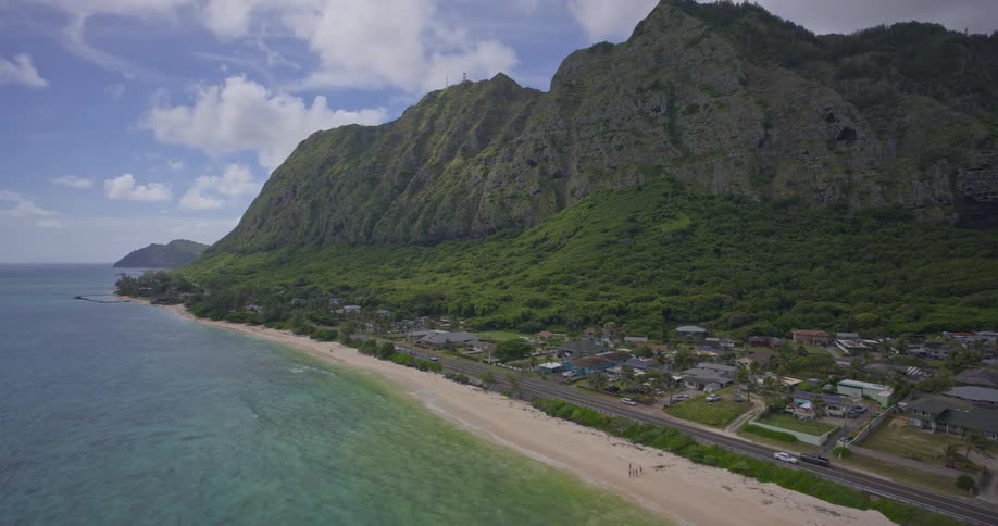 A slow aerial view reveals a picturesque coastal town nestled between a towering green mountain range and the beautiful turquoise Pacific Ocean