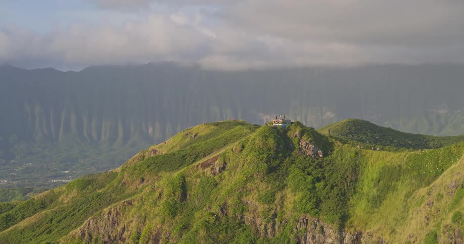 A stunning aerial view of hikers on top of a lush, green mountain ridge with a vast mountain range in the background
