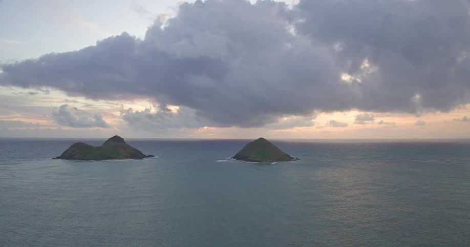 A tranquil wide shot of the two Mokulua Islands in the Pacific Ocean under a dramatic, cloudy sunrise