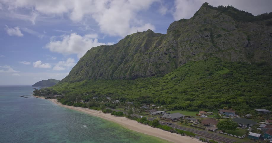 Static aerial view of a small coastal town nestled between a lush green mountain and the turquoise Pacific Ocean in Hawaii