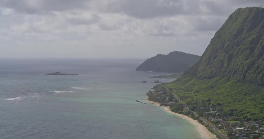 A scenic wide shot of a lush green mountain range meeting the ocean along a tropical coastline on a cloudy day