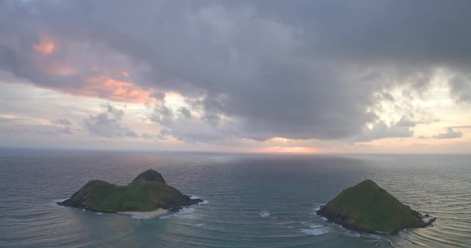 A serene, wide shot captures the Mokulua Islands at sunrise under a dramatic cloudy sky in Oahu, Hawaii