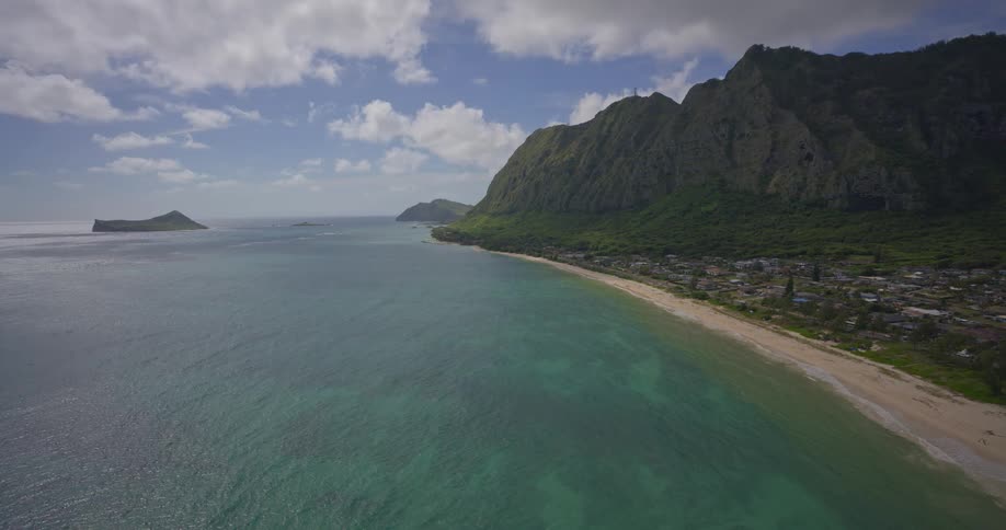 A serene, static aerial view shows the beautiful turquoise ocean meeting a sandy beach alongside a lush green mountain range in Hawaii
