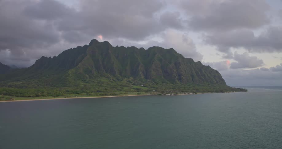 A serene, static wide shot captures the majestic green mountains of a tropical island coastline meeting the calm ocean under a cloudy sky