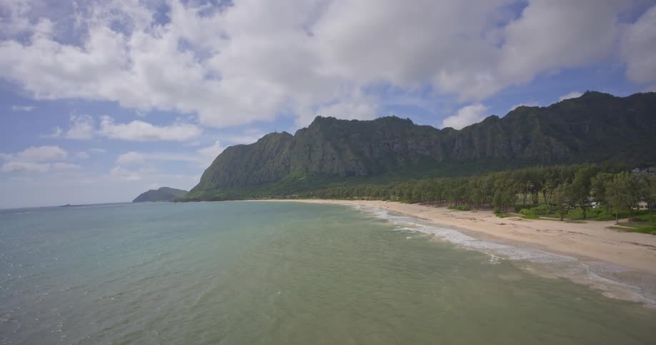 A stunning aerial view reveals a magnificent tropical coastline with lush green mountains, a pristine sandy beach, and a rolling ocean