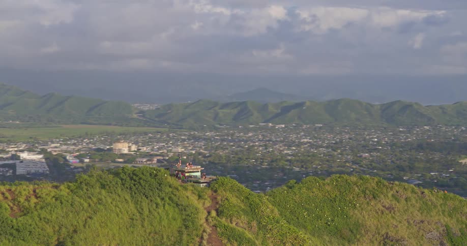 Sweeping aerial shot reveals a breathtaking panoramic view of a coastal city and bay from a historic pillbox on a lush green ridge in Hawaii