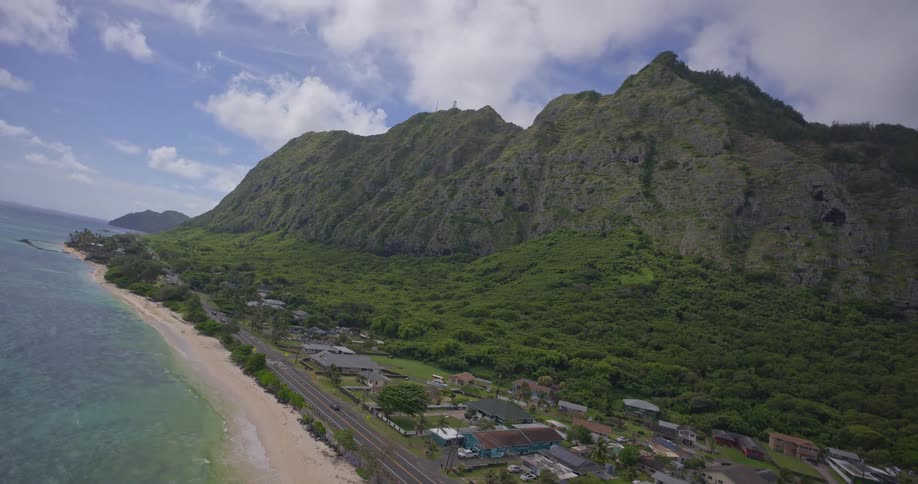 An expansive aerial view reveals a tropical coastline with a beach, a road, and a town nestled against a lush green mountain range
