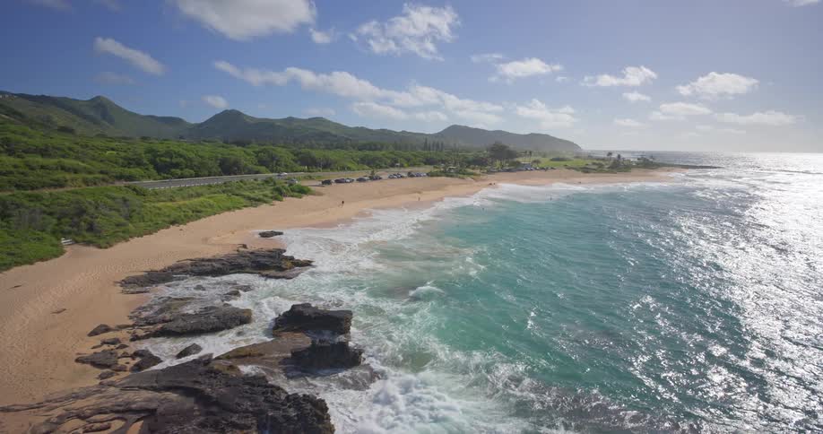A stunning aerial view of a tropical Hawaiian coastline with turquoise waves crashing on a sandy beach below green mountains