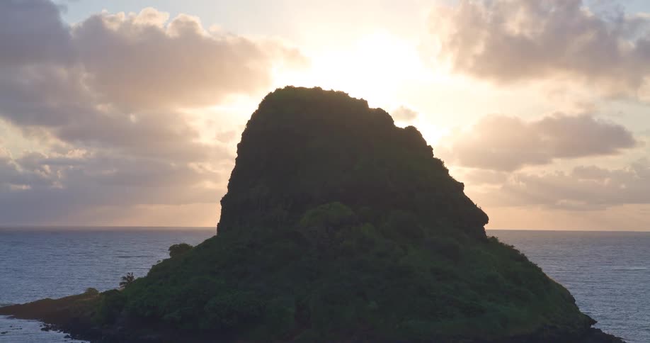 A golden sunrise peeks out from behind a lush tropical island silhouetted against the vast ocean