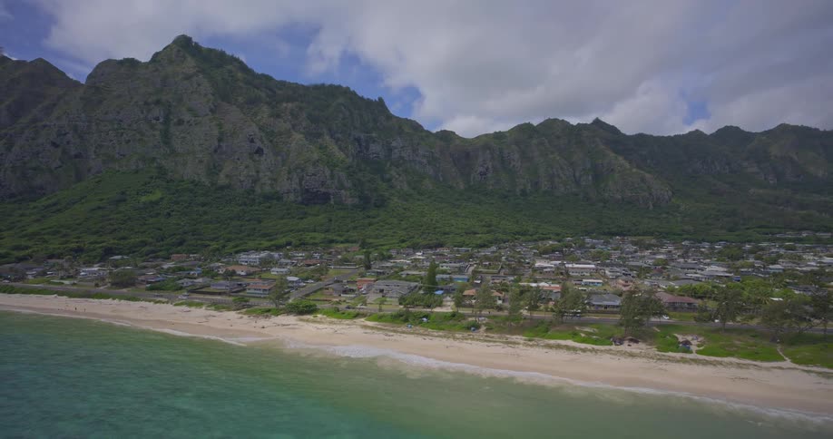 Aerial view pulling back over a tropical beach and ocean to reveal a coastal town nestled below dramatic green mountains
